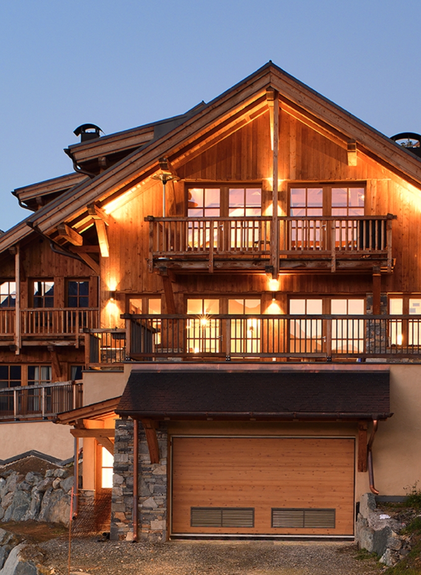 Vue de face d'un chalet illuminé ADVP, mise en valeur des 2 étages avec balcons et garage.